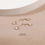 Four gold-plated barbell stud earrings with white pearl bezel settings, arranged on a beige textured surface with a white curved pedestal. Text overlay reads 'Gaugr: 18g Bar Length: 8mm'.