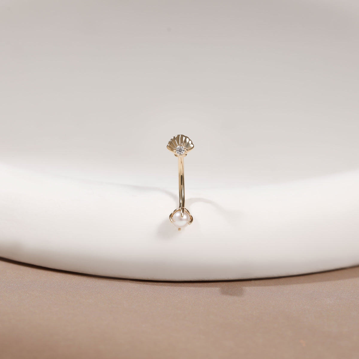 A single gold-toned navel piercing ring with a scalloped shell-shaped top and a clear faceted gemstone at the bottom, displayed on a white curved surface.