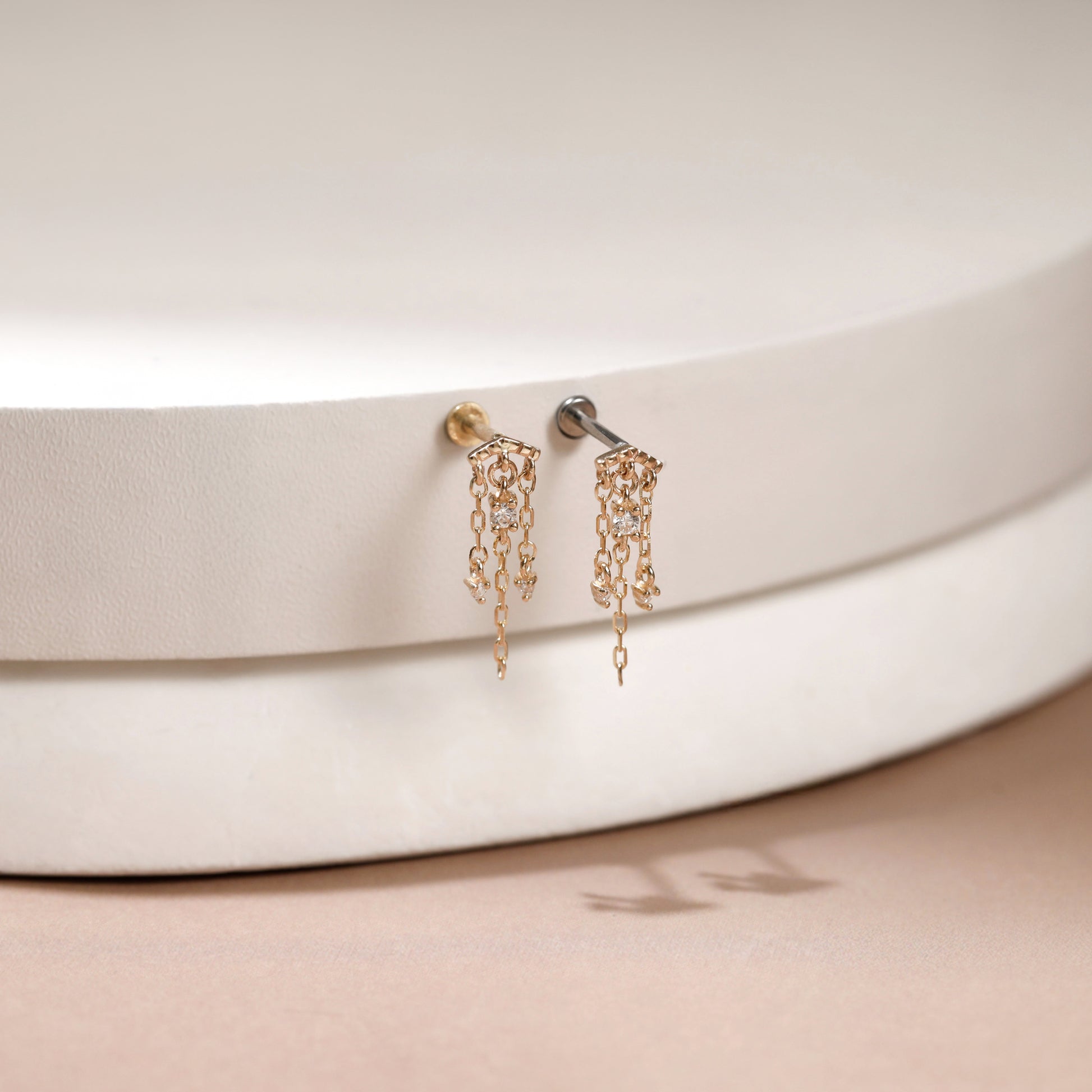 A pair of gold-toned dangle earrings with intricate chain-link designs and small hanging charms, displayed on a white textured circular stand against a soft beige background.