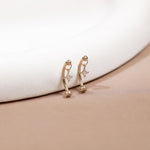 A pair of gold huggie hoop earrings with small star charms and tiny clear stones, resting on a beige surface next to a white curved display stand.