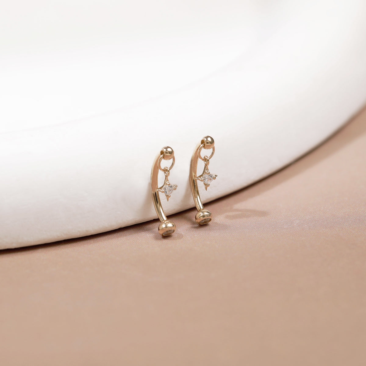 A pair of gold huggie hoop earrings with small star charms and tiny clear stones, resting on a beige surface next to a white curved display stand.