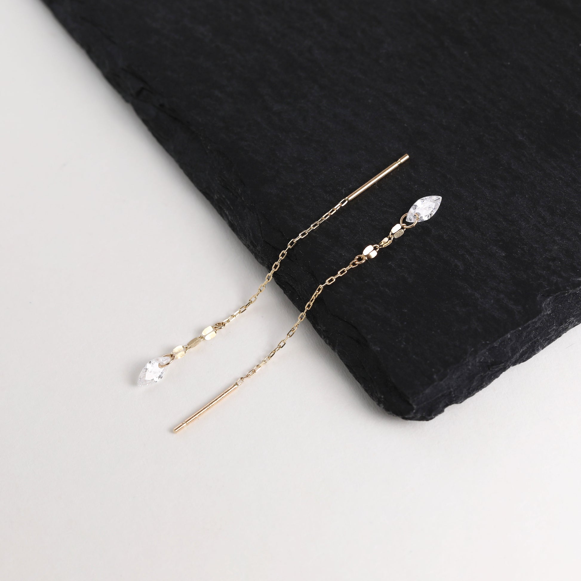 A pair of long gold-tone dangle earrings with clear faceted gemstones, displayed on a black slate slab over a light gray background.