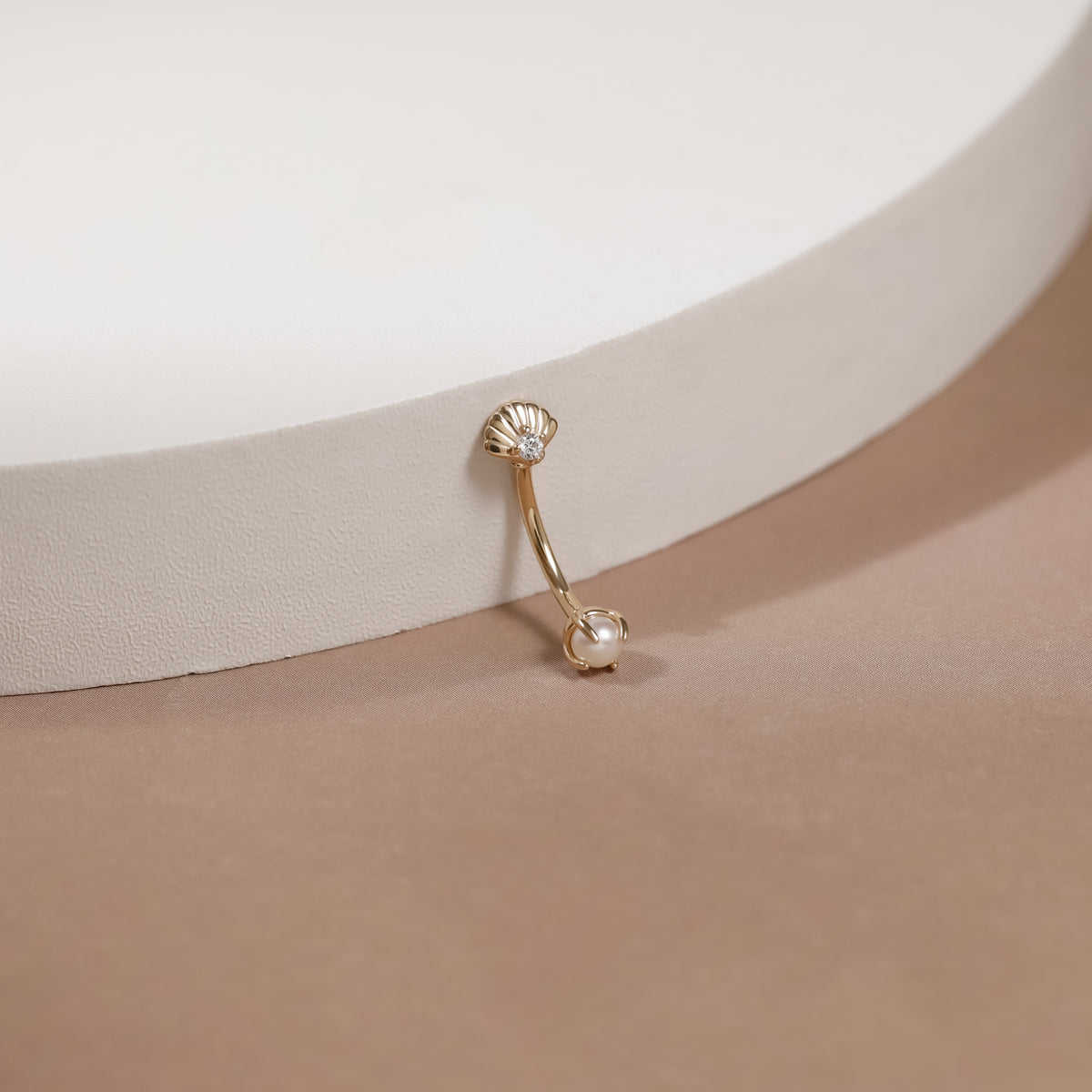 A gold curved barbell belly button ring with a small clear stone on the top and a white pearl on the bottom, displayed on a beige surface next to a white curved display block.