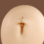 A close-up of a small gold navel piercing stud with a ring, a tiny clear star-shaped crystal, and a dangling clear bead, shown on a beige mannequin torso against a brown background.