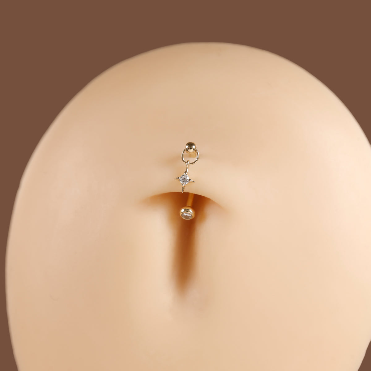 A close-up of a small gold navel piercing stud with a ring, a tiny clear star-shaped crystal, and a dangling clear bead, shown on a beige mannequin torso against a brown background.