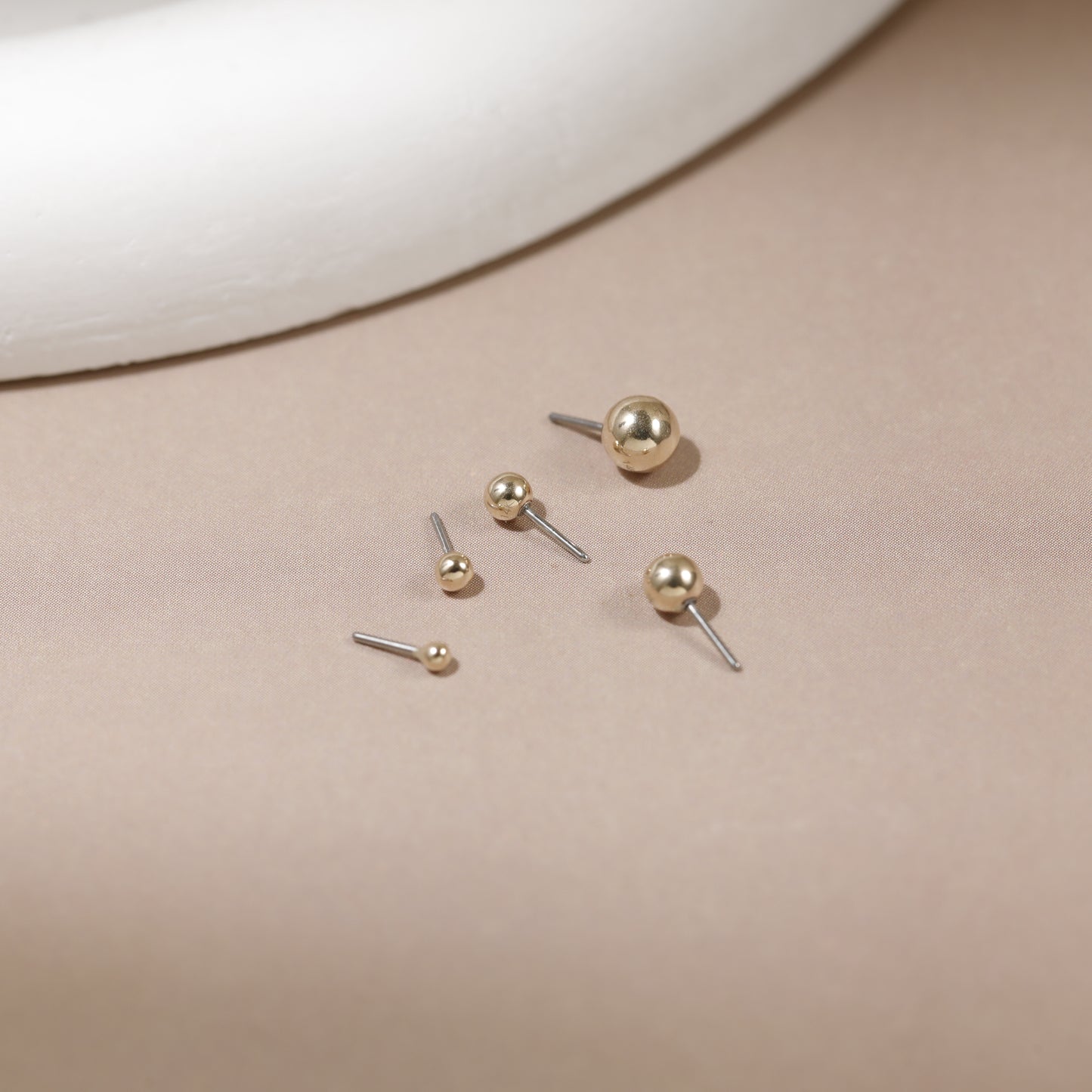 Five gold ball stud earrings in various sizes with silver-toned posts, arranged on a beige surface with a white curved prop in the background.