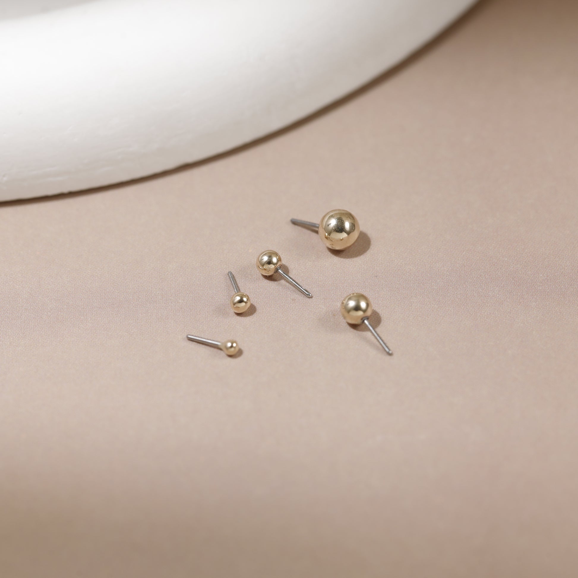 Five gold ball stud earrings in various sizes with silver-toned posts, arranged on a beige surface with a white curved prop in the background.