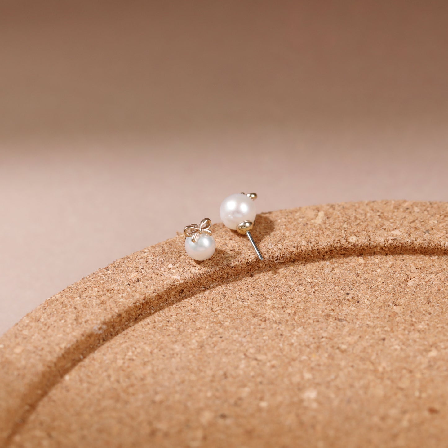 A pair of small stud earrings with white pearl-like beads and silver-toned posts, resting on a curved tan textured surface against a soft beige background.