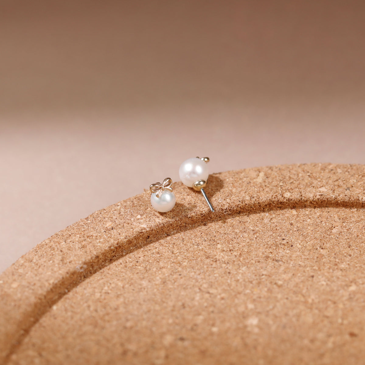 A pair of small stud earrings with white pearl-like beads and silver-toned posts, resting on a curved tan textured surface against a soft beige background.