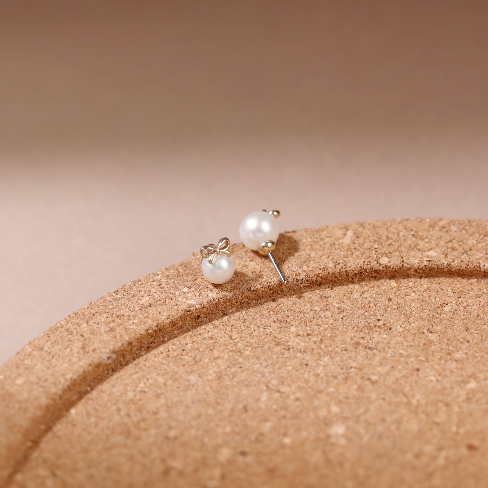 A pair of small stud earrings with white pearl-like beads and silver-toned posts, resting on a curved tan textured surface against a soft beige background.