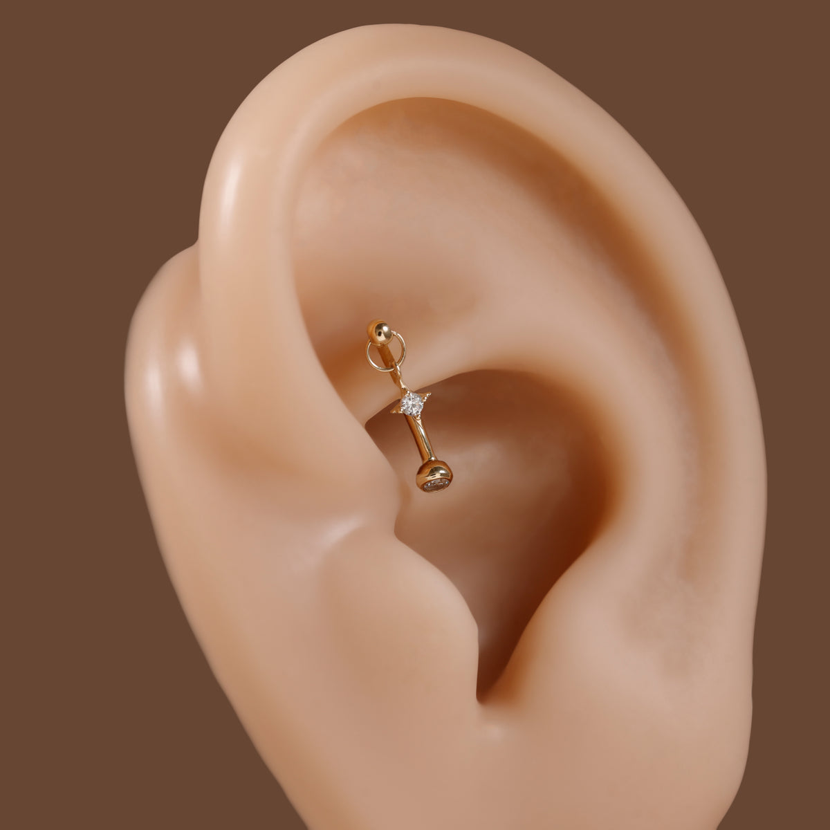 A gold ear cartilage piercing stud with a small clear crystal accent, shown on a beige silicone ear model against a solid brown background.
