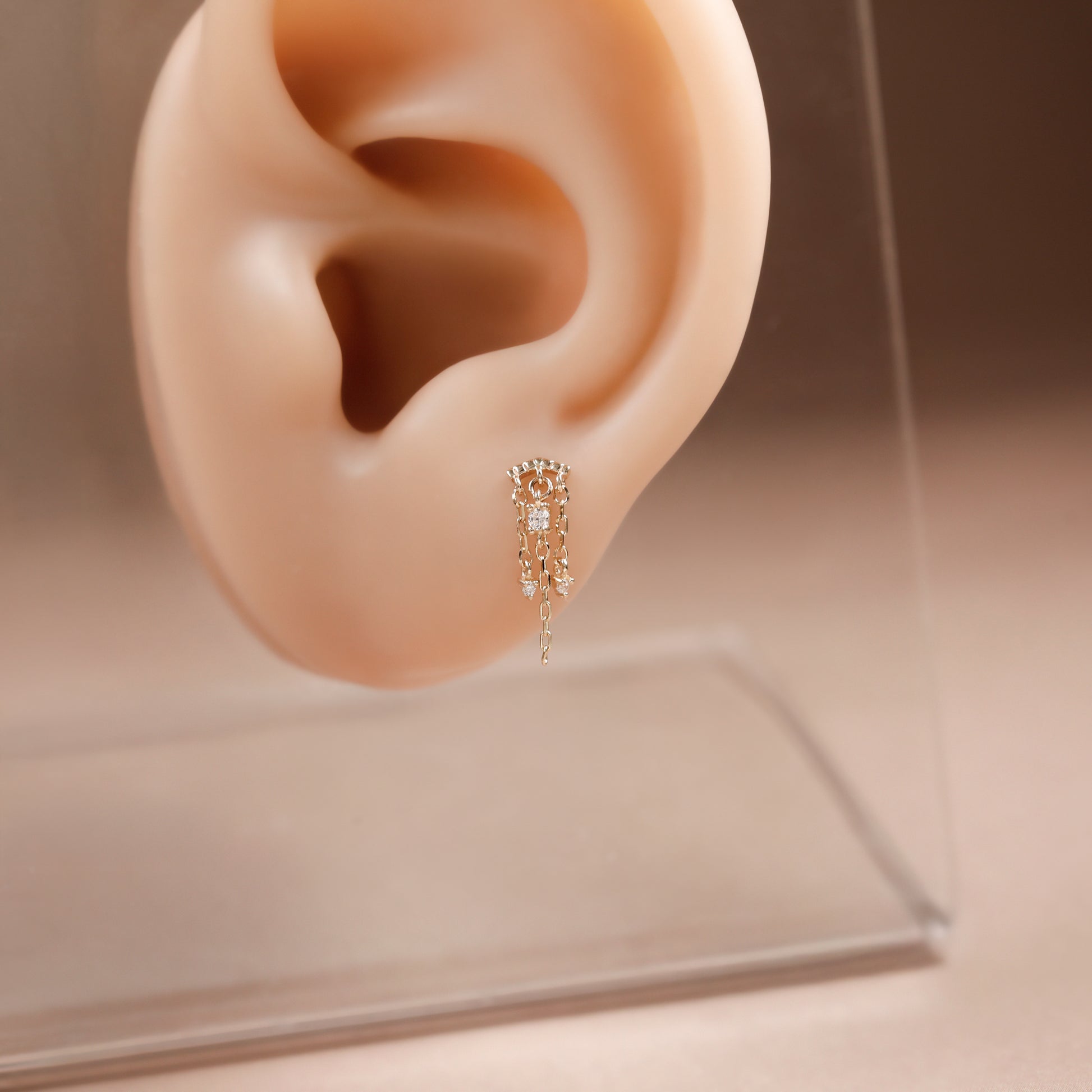 A delicate gold dangle earring with clear gemstones is displayed on a beige silicone ear model stand, set against a soft beige background with a clear acrylic display base.