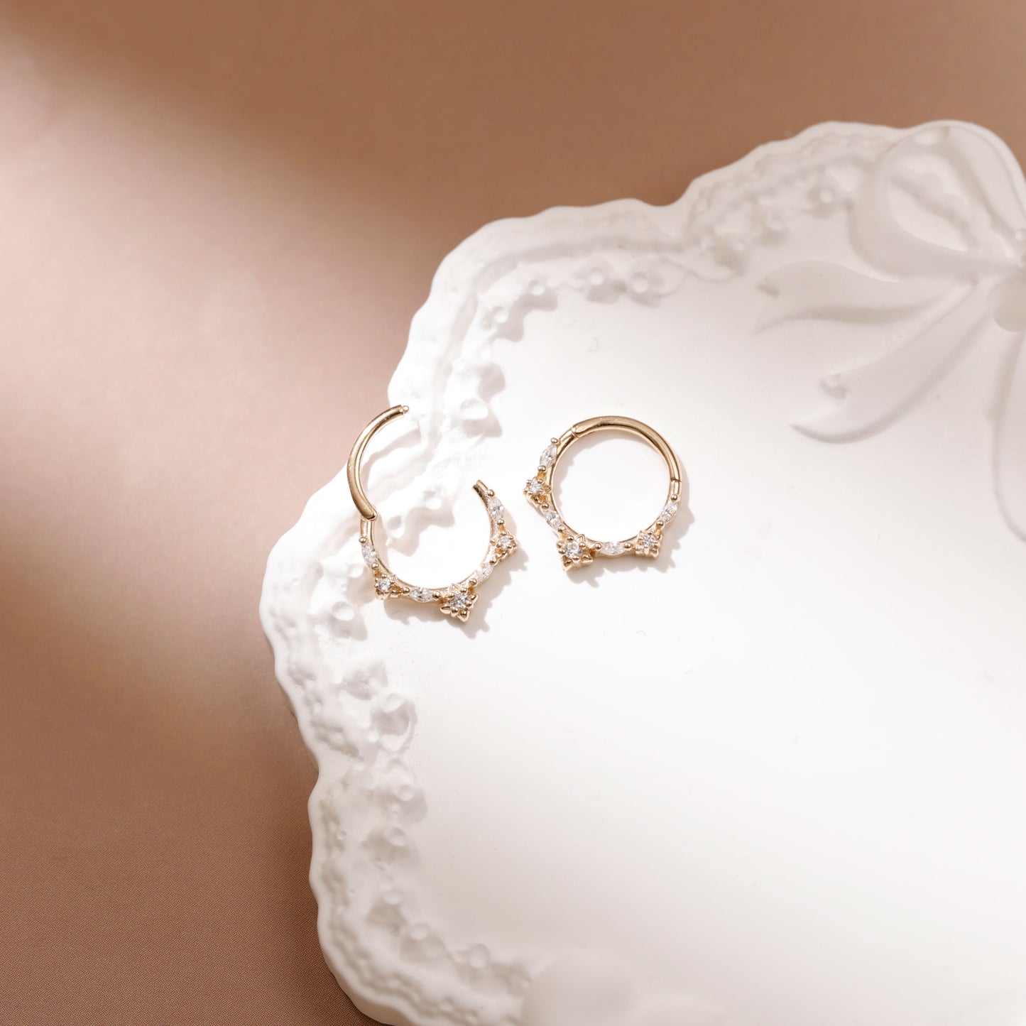 A pair of gold crescent moon hoop earrings adorned with small clear crystals, displayed on a white decorative ceramic plate against a warm beige background.