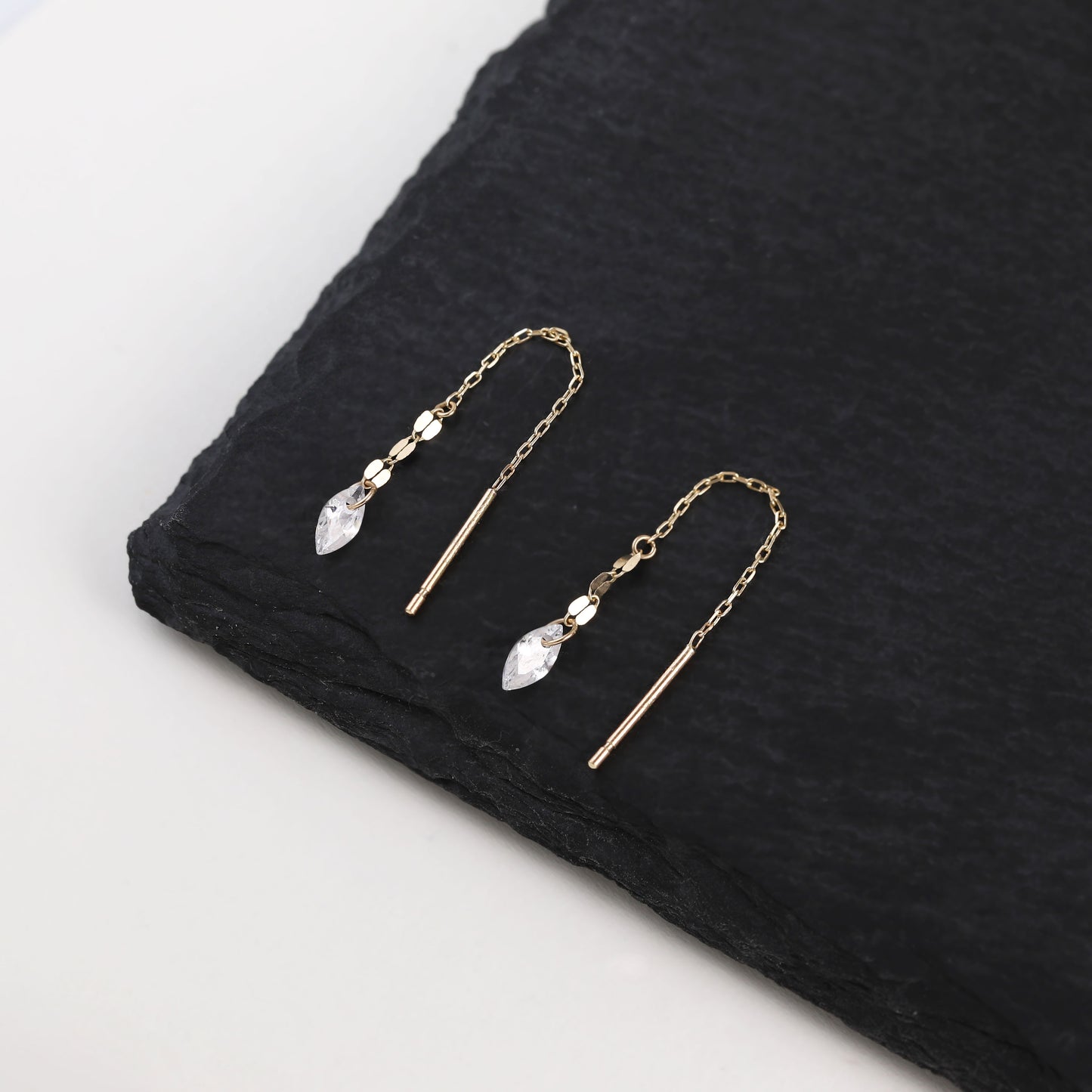A pair of gold-toned drop earrings with clear faceted teardrop crystals and small clear beads, displayed on a black textured stone slab against a white background.
