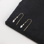 A pair of gold-toned drop earrings with clear faceted teardrop crystals and small clear beads, displayed on a black textured stone slab against a white background.