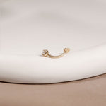 A gold curved barbell piercing stud with a small white pearl on one end, resting on a white ceramic plate against a beige background.