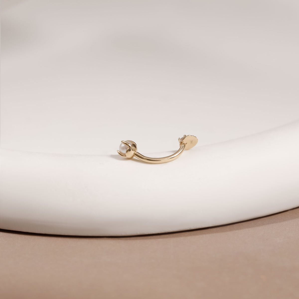 A gold curved barbell piercing stud with a small white pearl on one end, resting on a white ceramic plate against a beige background.
