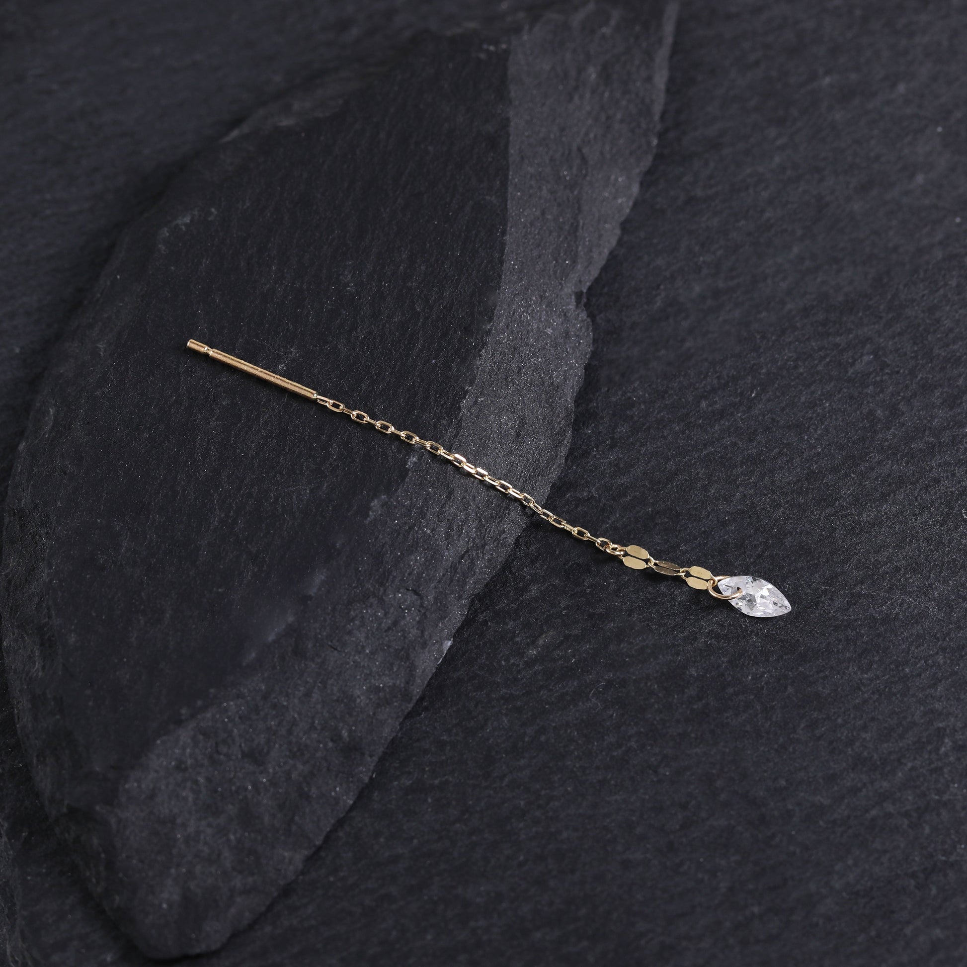 A delicate gold hair pin with a small clear marquise-cut crystal charm, displayed on a dark textured slate surface.