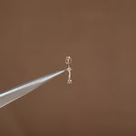 A tiny gold earring with a small clear gemstone, held delicately between metal tweezers against a solid brown background.