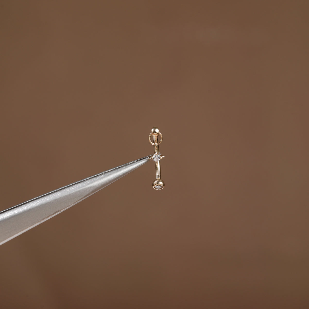 A tiny gold earring with a small clear gemstone, held delicately between metal tweezers against a solid brown background.