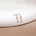 A pair of gold hoop earrings with small dangling star charms, displayed on a white curved surface with a soft beige background.