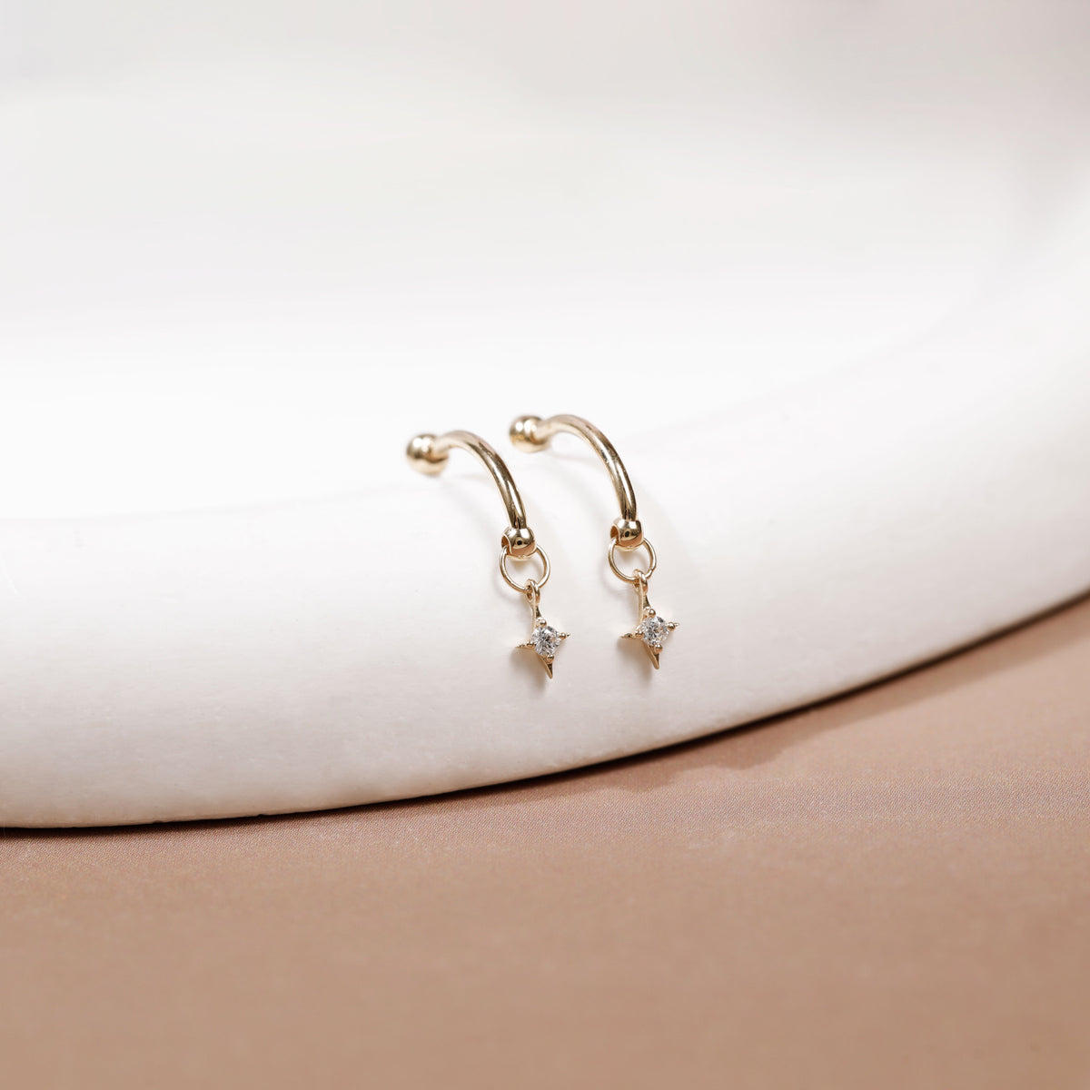 A pair of gold hoop earrings with small dangling star charms, displayed on a white curved surface with a soft beige background.