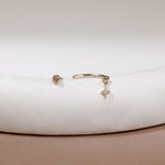A pair of gold body jewelry, including a curved barbell stud earring with a small dangling crystal star charm and a matching round stud earring, displayed on a white curved jewelry stand.