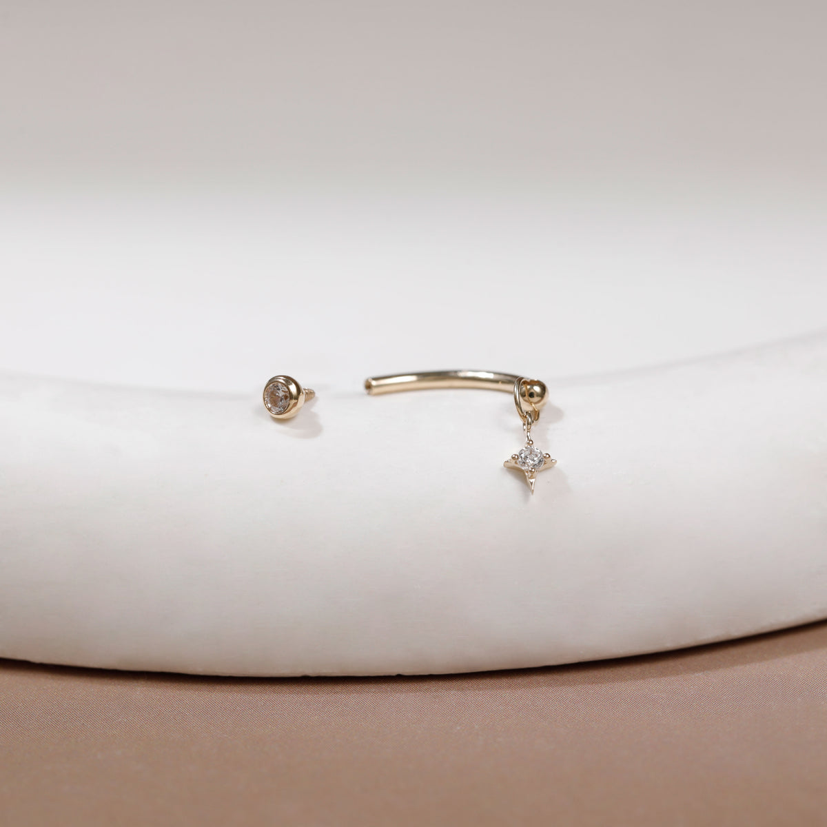 A pair of gold body jewelry, including a curved barbell stud earring with a small dangling crystal star charm and a matching round stud earring, displayed on a white curved jewelry stand.