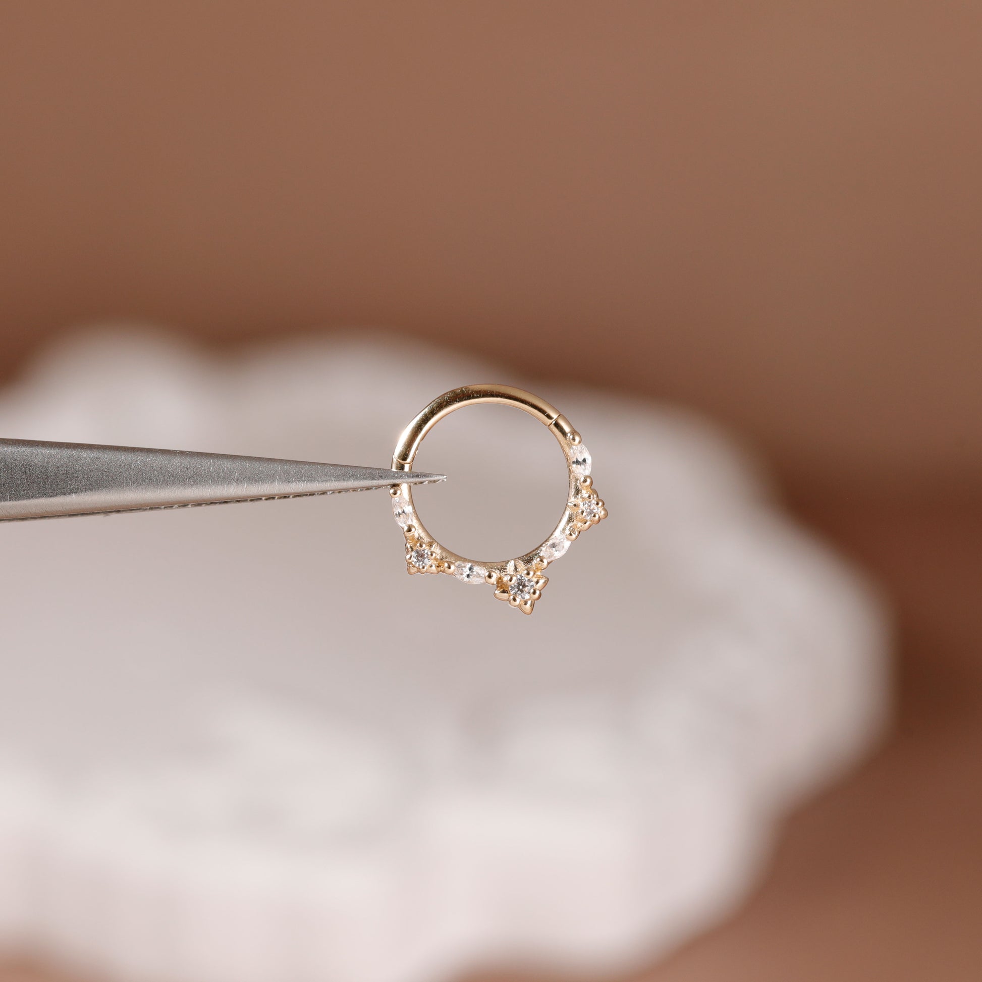 A delicate gold ring with small clear stones, held by metal tweezers against a soft, blurred brown and white background.