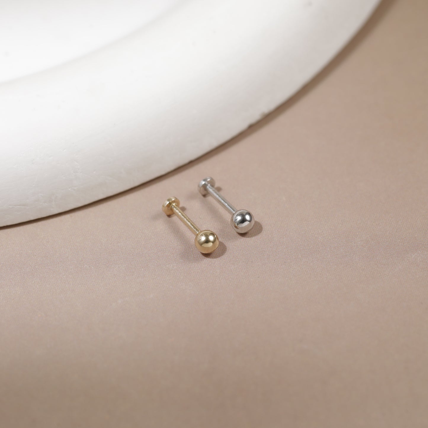Two small barbell stud earrings, one gold-toned and one silver-toned, displayed on a beige surface with a white curved prop in the background.