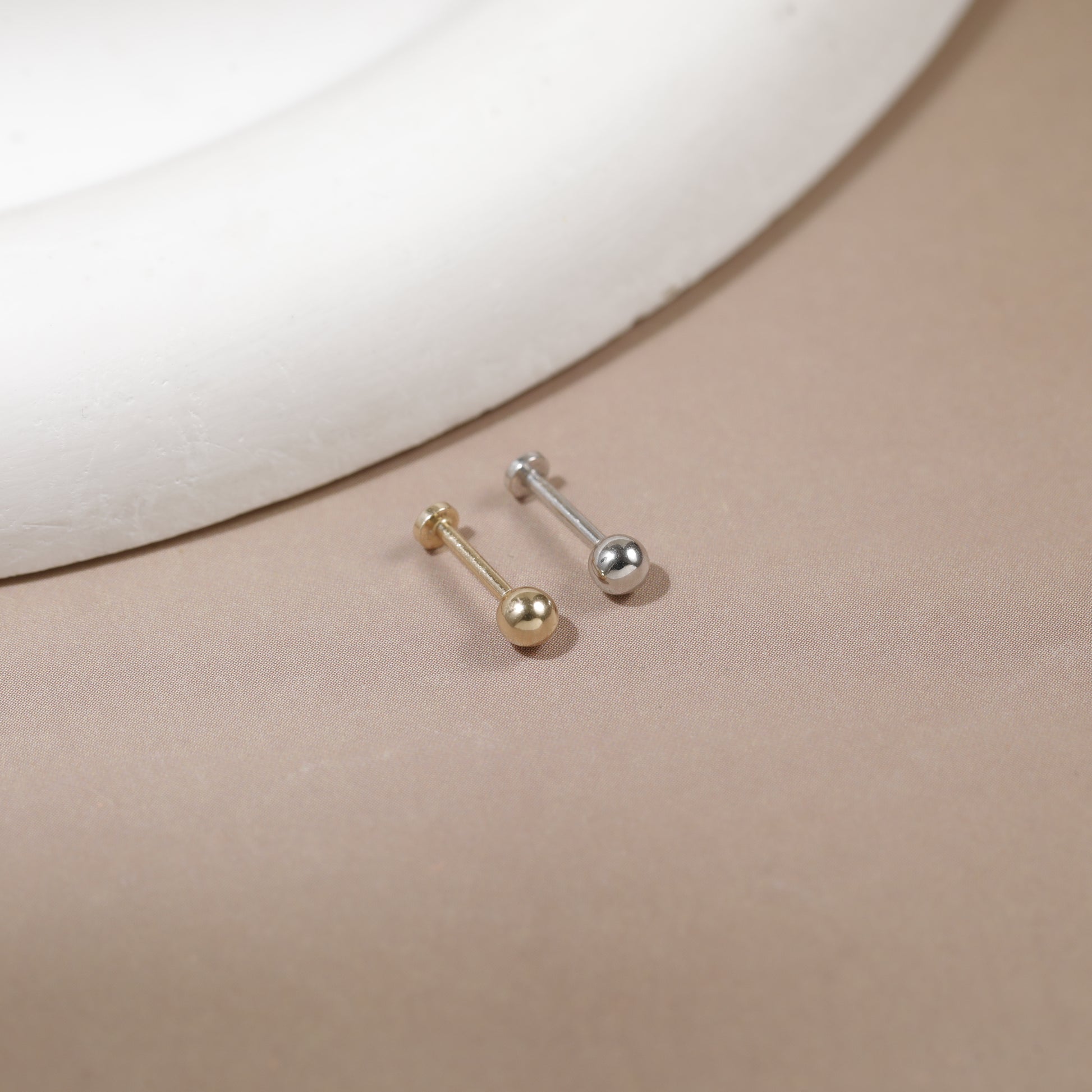 Two small barbell stud earrings, one gold-toned and one silver-toned, displayed on a beige surface with a white curved prop in the background.