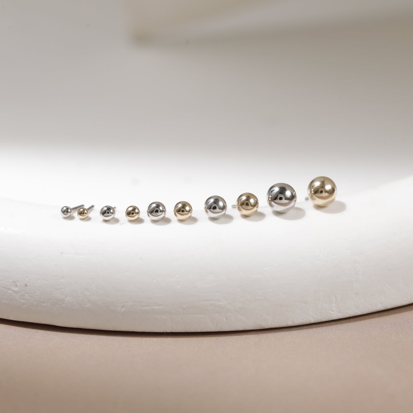 A set of nine spherical stud earrings in graduated sizes, featuring a mix of silver and gold finishes, displayed on a curved white surface.