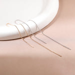 Three long drop earrings in rose gold, yellow gold, and silver, arranged on a beige surface with a white curved ceramic prop.