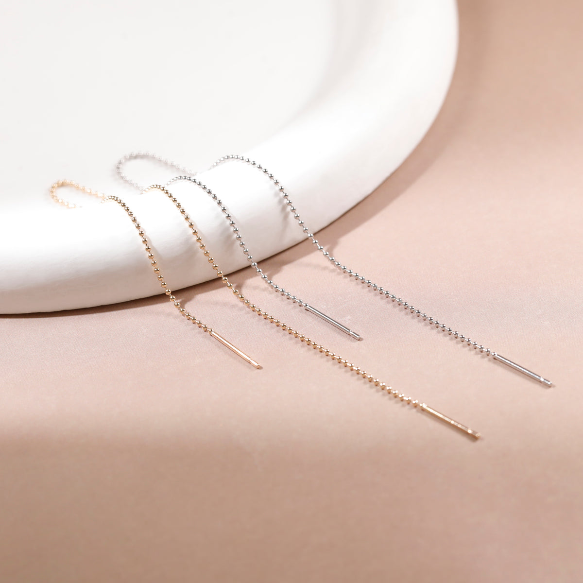 Three long drop earrings in rose gold, yellow gold, and silver, arranged on a beige surface with a white curved ceramic prop.
