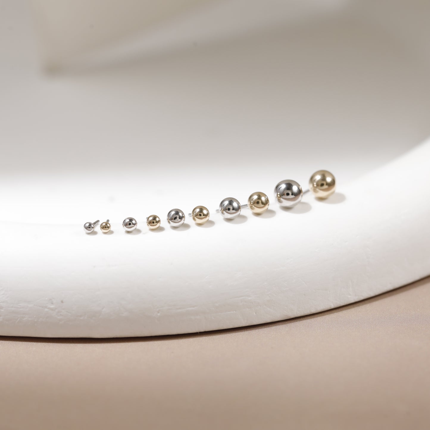 A set of nine small spherical stud earrings arranged in ascending size order on a white display form. The earrings are metallic, featuring alternating silver and gold finishes.