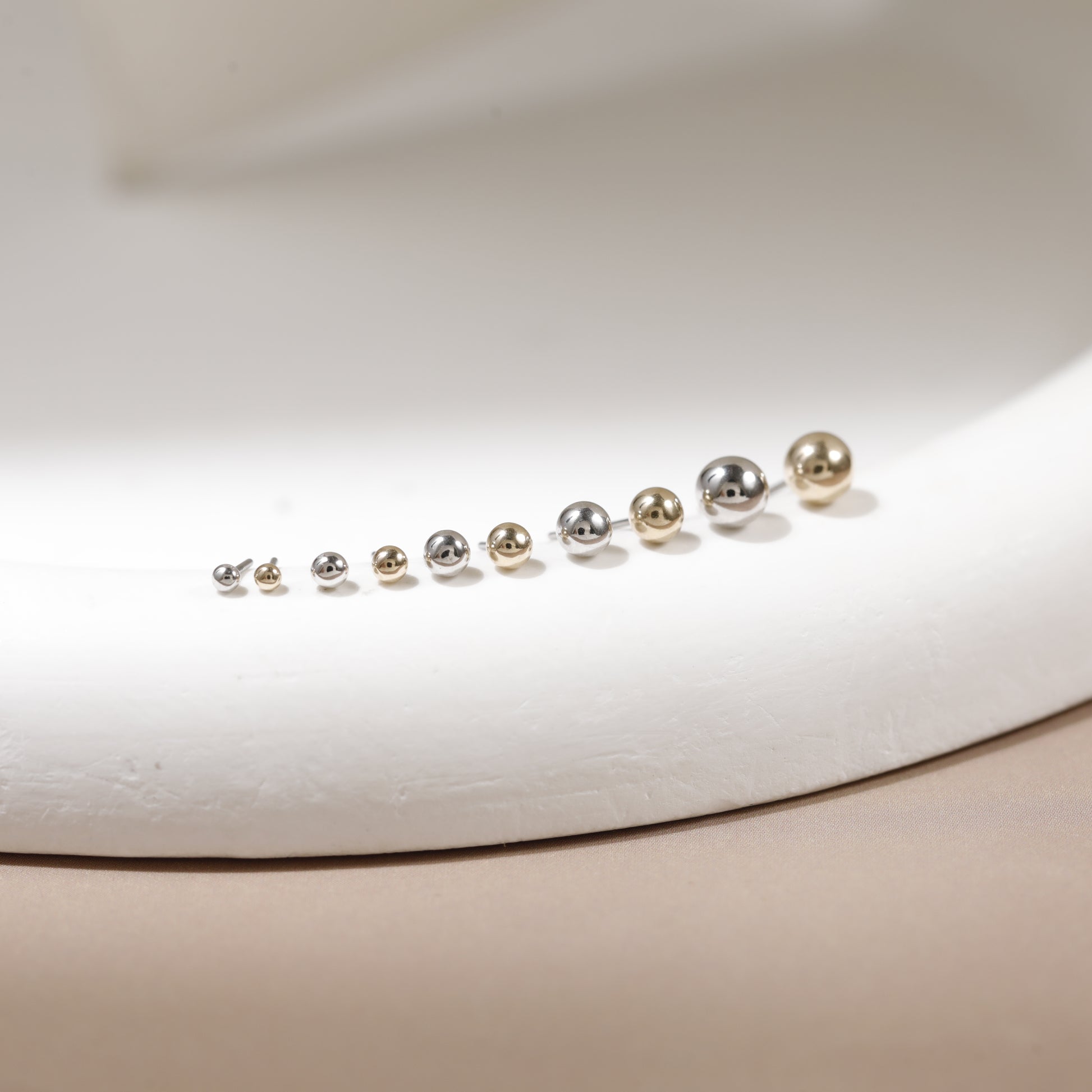 A set of nine small spherical stud earrings arranged in ascending size order on a white display form. The earrings are metallic, featuring alternating silver and gold finishes.