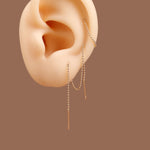 A pair of delicate gold chain dangle earrings with small bead links, shown on a beige ear mannequin against a solid brown background.