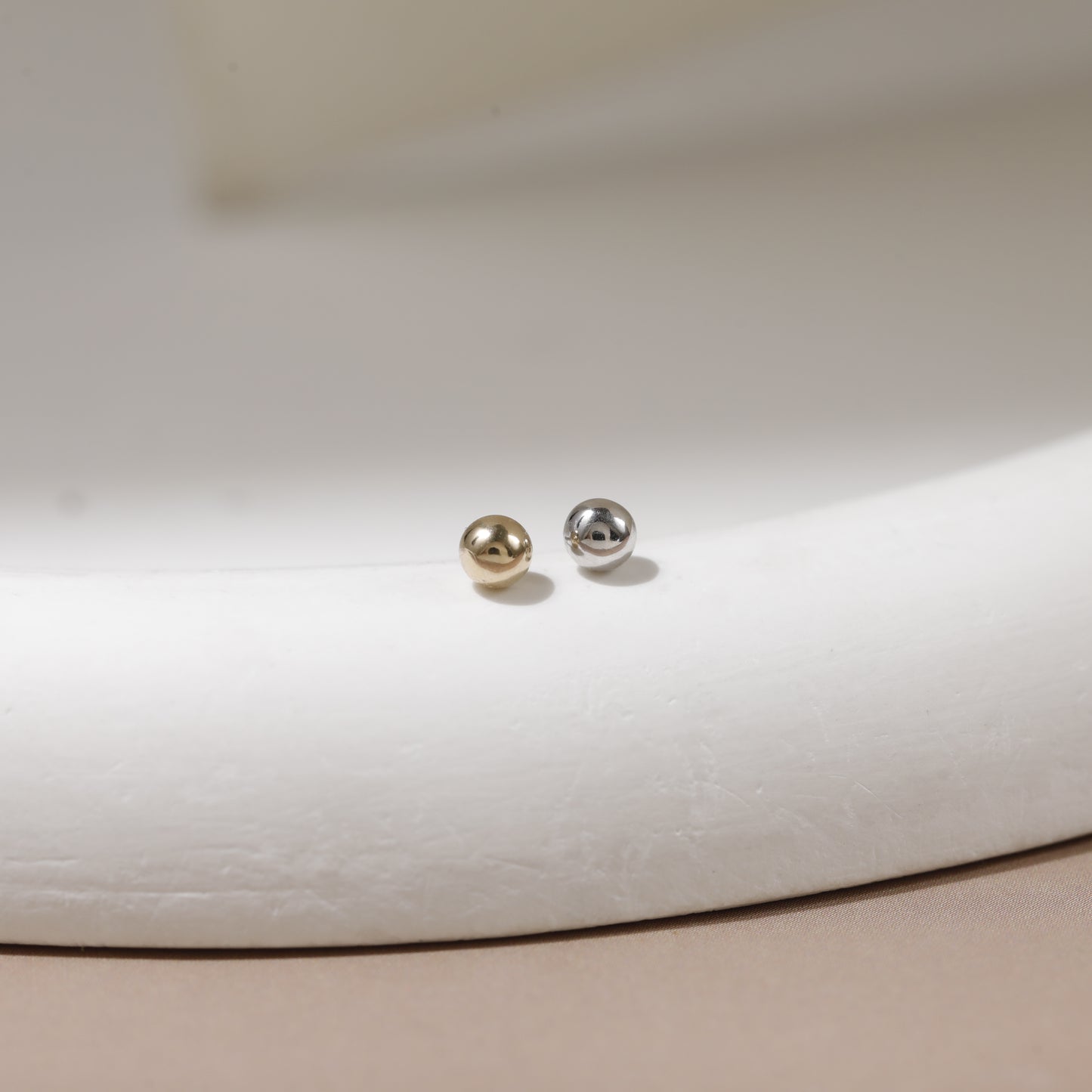 A pair of small spherical stud earrings, one in polished gold and one in polished silver, displayed on a curved white jewelry stand.