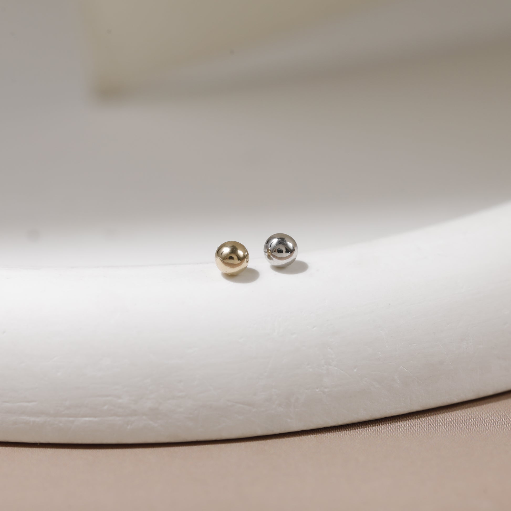A pair of small spherical stud earrings, one in polished gold and one in polished silver, displayed on a curved white jewelry stand.