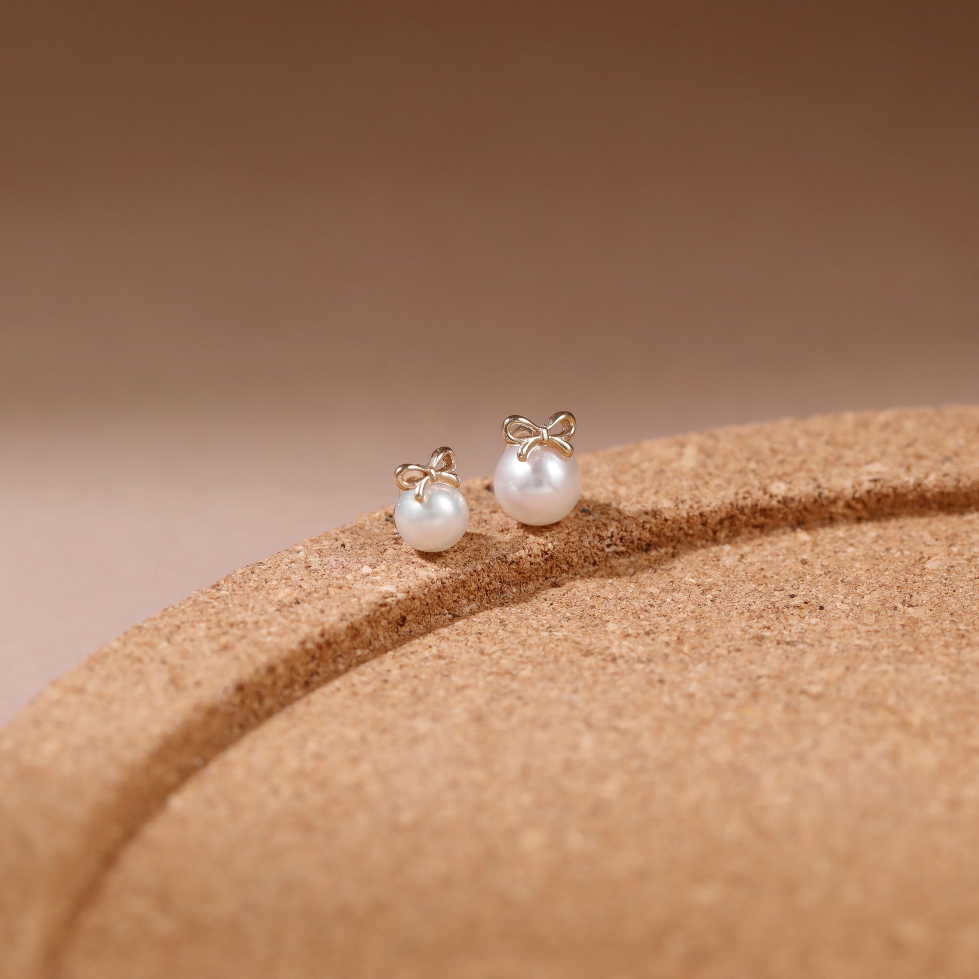 A pair of gold bow stud earrings with white pearl-like beads, resting on a textured brown display stand against a warm beige background.