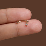 A close-up of a tiny gold curved barbell stud earring with a dangling clear gemstone, shown resting on a person's fingers against a solid brown background.