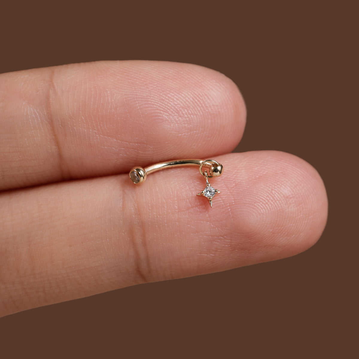 A close-up of a tiny gold curved barbell stud earring with a dangling clear gemstone, shown resting on a person's fingers against a solid brown background.