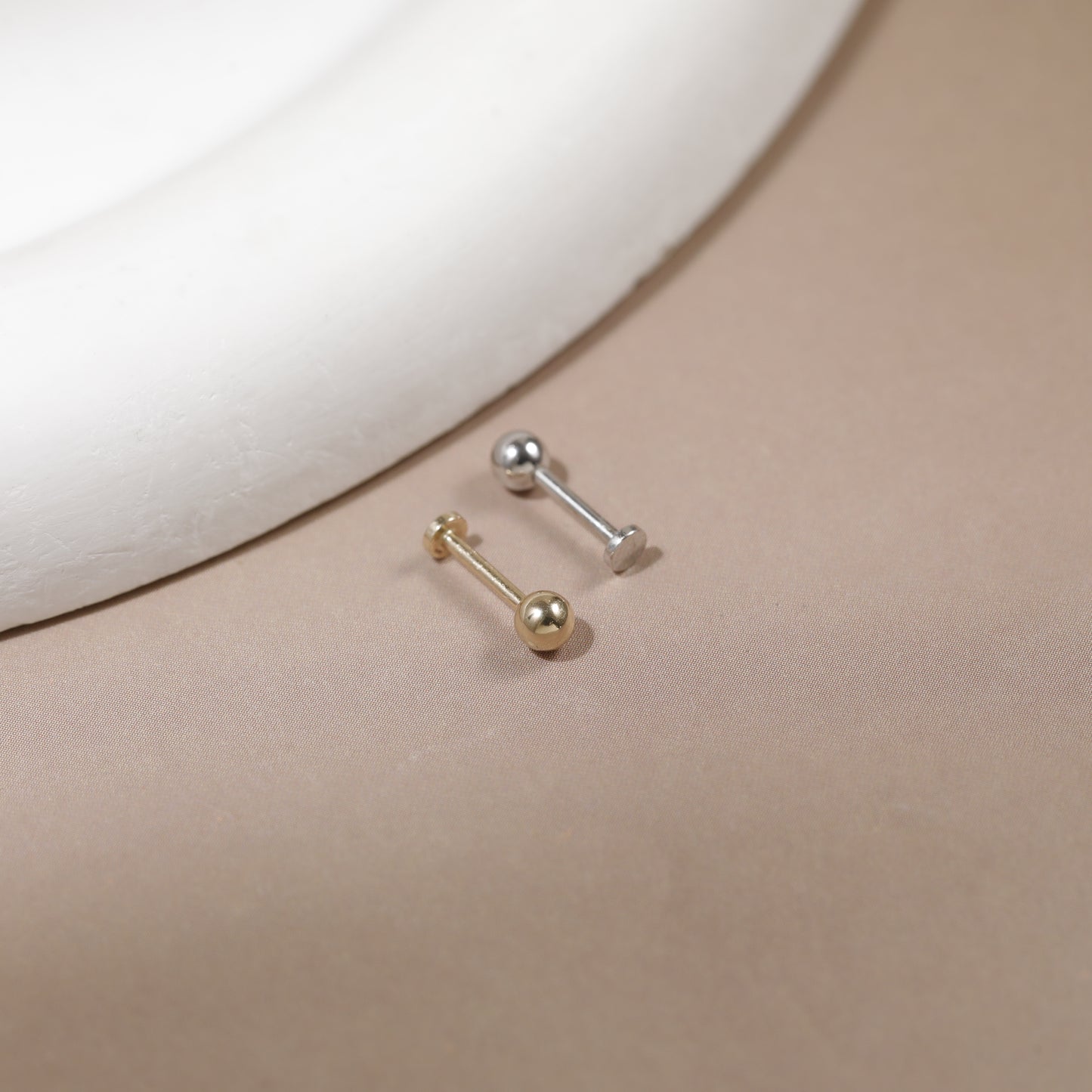 Two small barbell stud earrings, one gold-toned and one silver-toned, displayed on a beige surface next to a white curved display prop.
