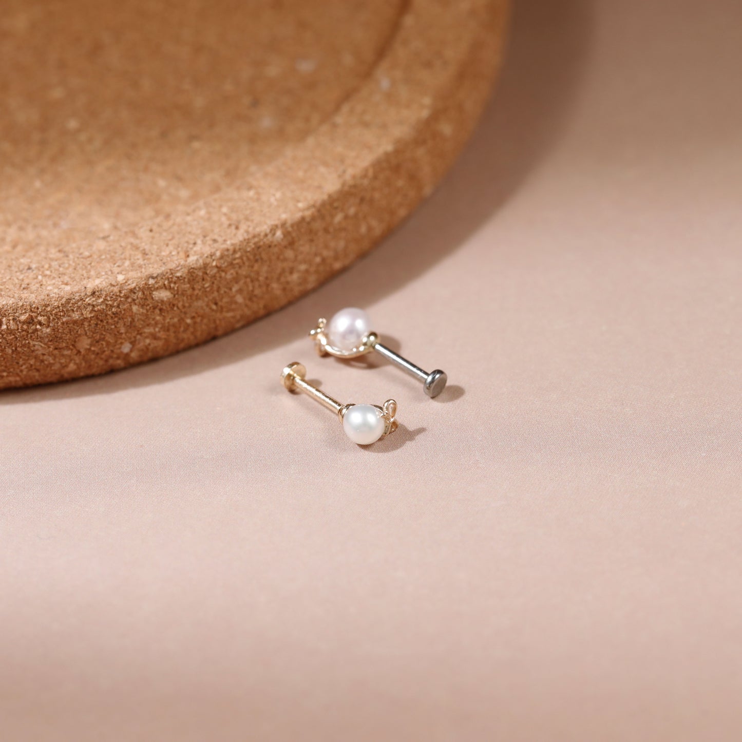 A pair of gold and silver barbell stud earrings, each adorned with a small white pearl, displayed on a soft beige surface next to a cork coaster.