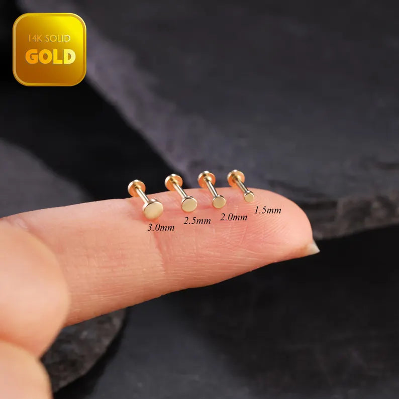Gold nose studs of different sizes displayed on a finger with a dark background.