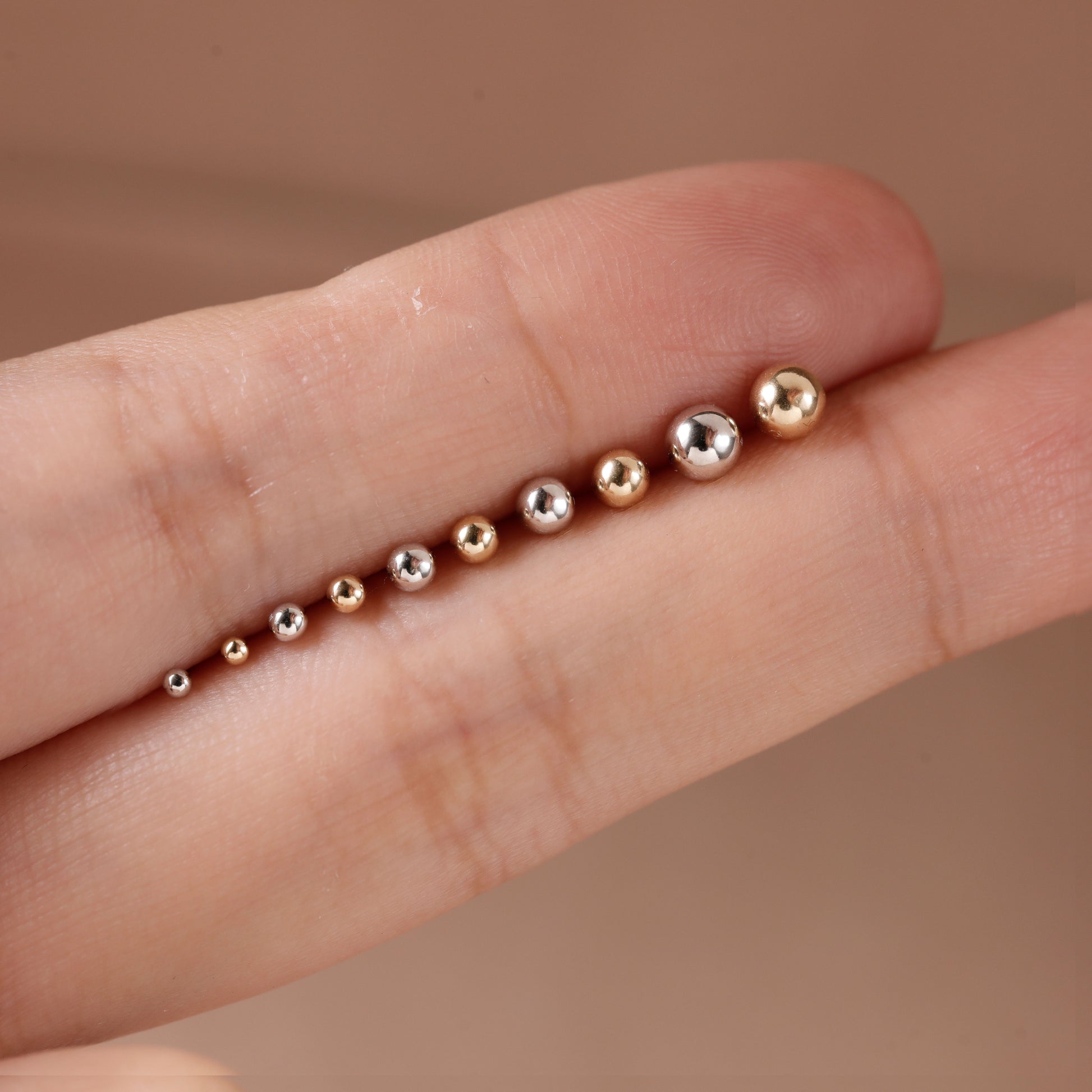 A close-up of a person's finger displaying a row of small metallic beads in graduated sizes, including rose gold, yellow gold, and silver.