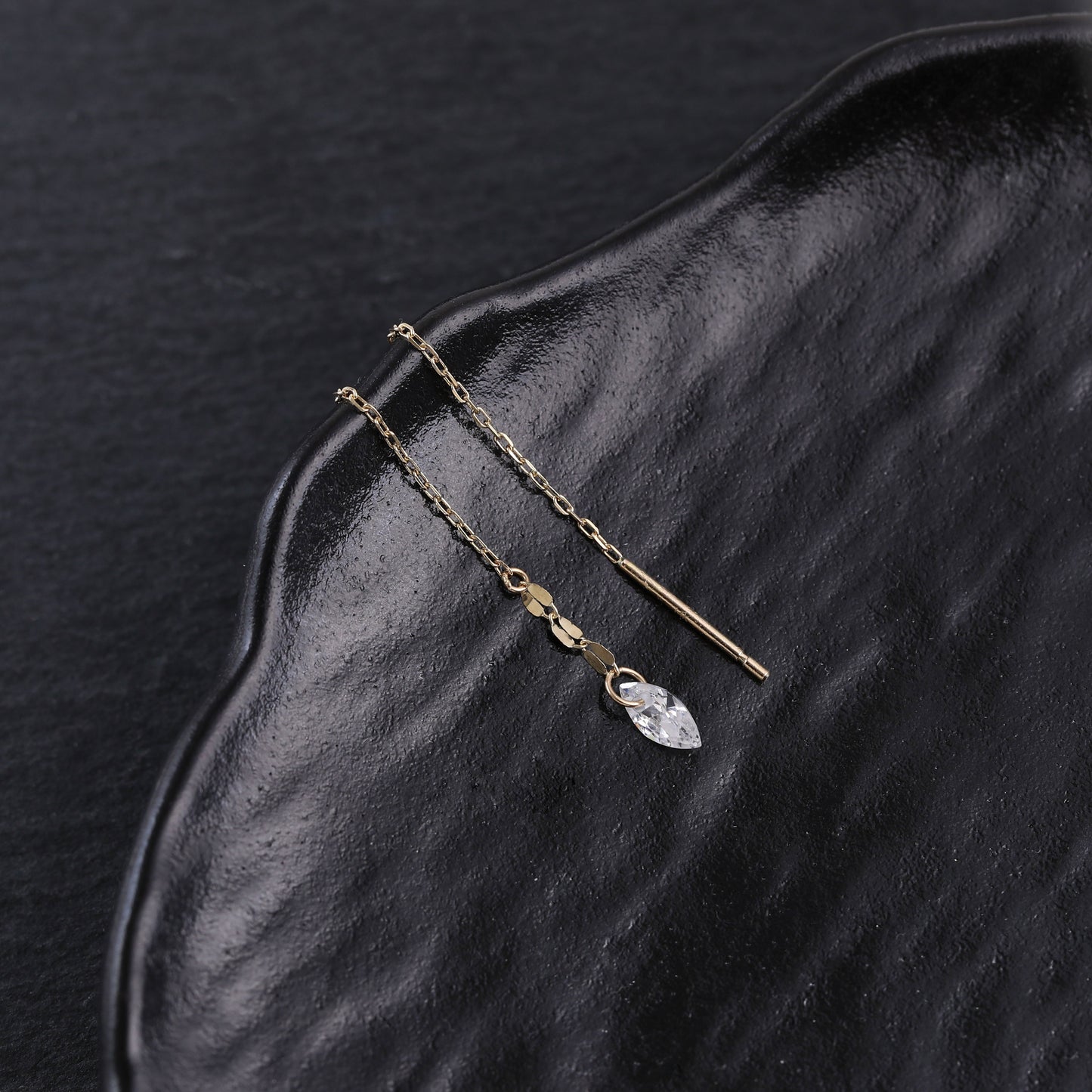 A pair of gold-toned drop earrings with a small clear faceted crystal pendant, displayed on a textured black surface.