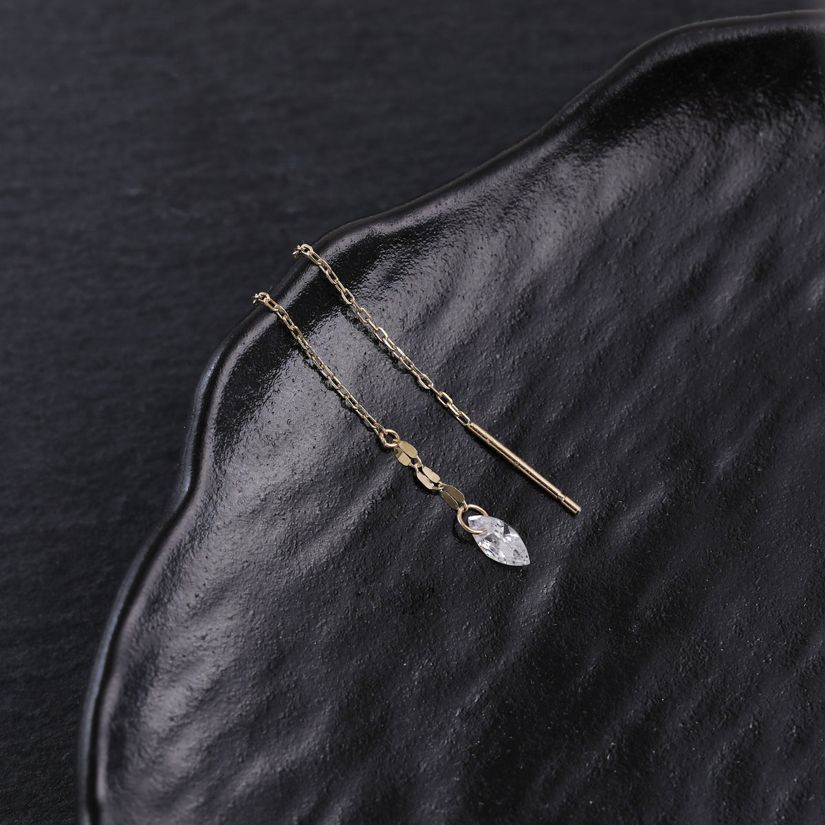 A pair of gold-toned drop earrings with a small clear faceted crystal pendant, displayed on a textured black surface.