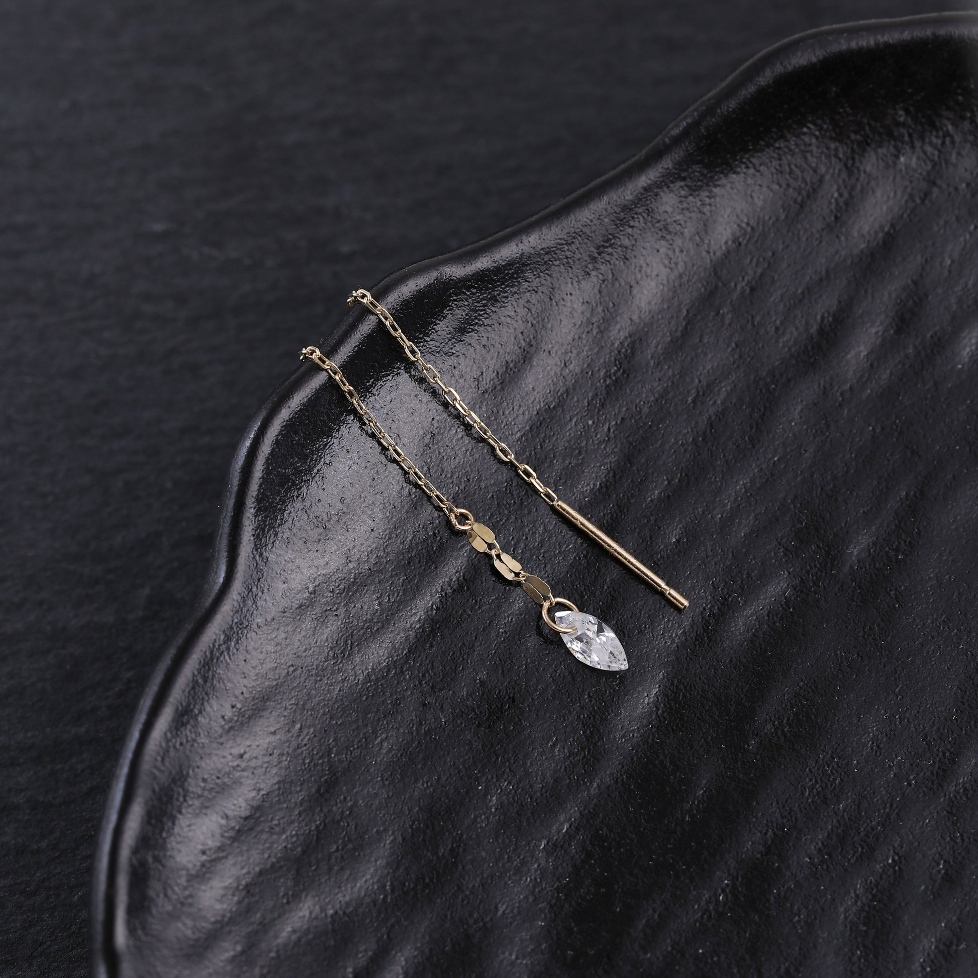 A pair of gold-toned drop earrings with a small clear faceted crystal pendant, displayed on a textured black surface.