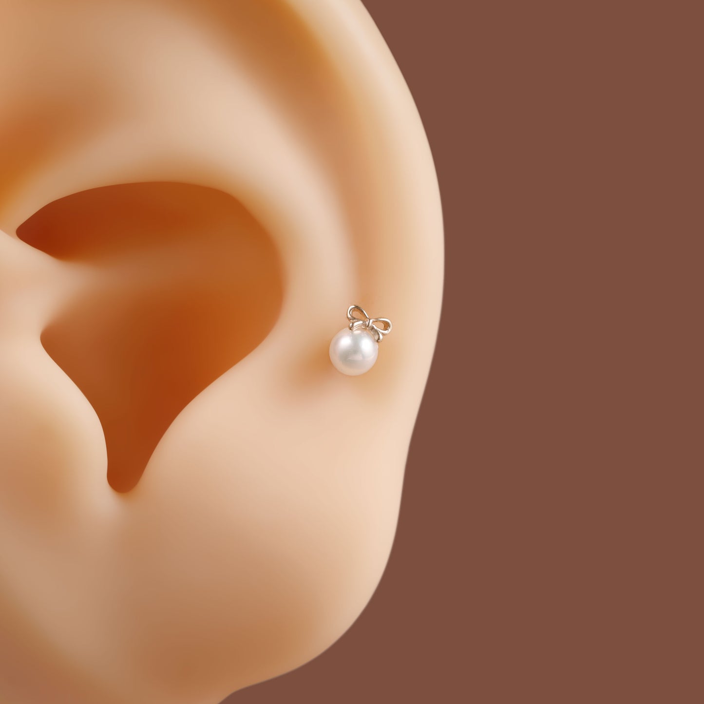 A single pearl stud earring with a small silver bow setting, shown worn on a beige ear against a solid brown background.